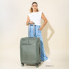 Front view of VersaPack®+ Medium Check-In Spinner in Desert Sage Green, shown with a 5'7" model for scale, highlighting the extended handle and upright rolling design