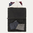 A black Travelpro Essentials washable laundry bag with a large zippered opening, displaying a white shirt and folded plaid and striped garments inside, alongside a mesh pocket with additional items.