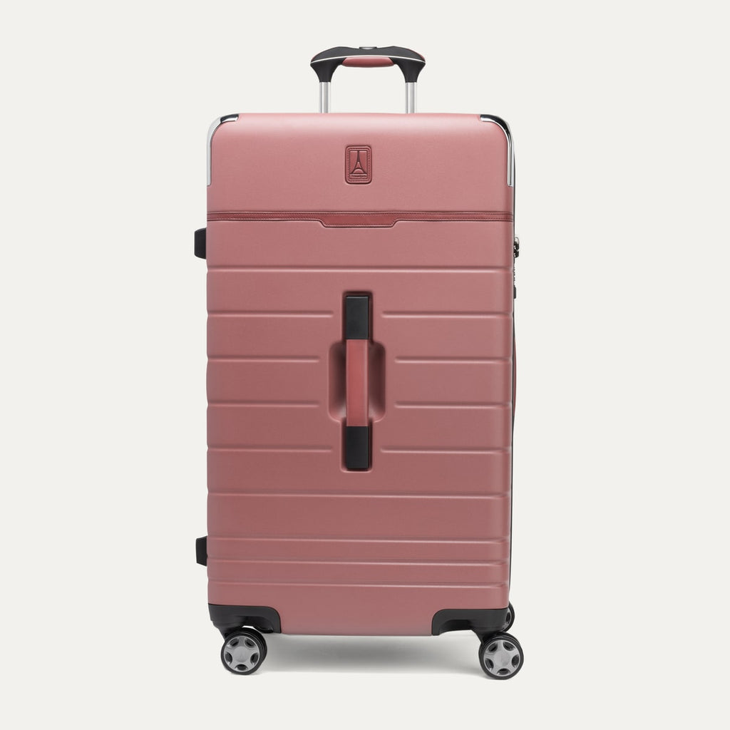 front view of the large check-in trunk spinner in astro dust red, showcasing its durable textured polycarbonate hardshell construction, signature zinc corner armor, premium brown leather trim accents, and four-wheeled spinner system with an extended telescoping handle.