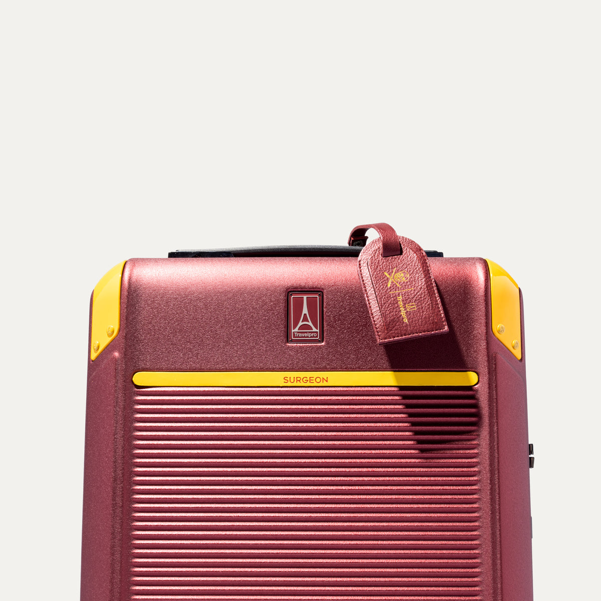 Top front detail of Travelpro x Surgeon carry on Spinner in Daring Red, featuring yellow trim, co-branded logo, and signature luggage tag.
