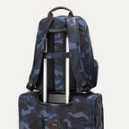 **Alt text:** Rear view of the Platinum® Elite Business Backpack in blue camouflage, secured to a matching suitcase handle using its built-in luggage pass-through strap.
