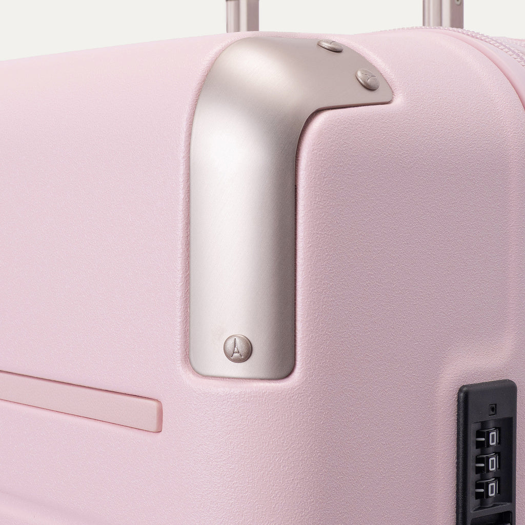 Close-up of the Travelpro Platinum Elite Medium Check-In Hardside Spinner in Powder Pink, featuring protective polished aluminum corner armor, an integrated TSA-approved combination lock, and smooth side zipper trim against a clean off-white background.