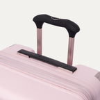 Close-up of the Travelpro Platinum Elite Medium Check-In Hardside Spinner in Powder Pink, highlighting the multi-stop PowerScope Lite telescoping handle, the rubberized Contour Grip, and the top carry handle against a clean off-white background.