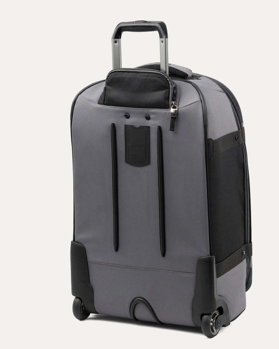 rear three-quarter view of the gray black bold by travelpro medium check-in expandable rollaboard suitcase standing vertically, showing its protective skid guards, molded corner guards, reinforced wheel housings, two high-performance ball-bearing wheels, and extended telescoping handle.