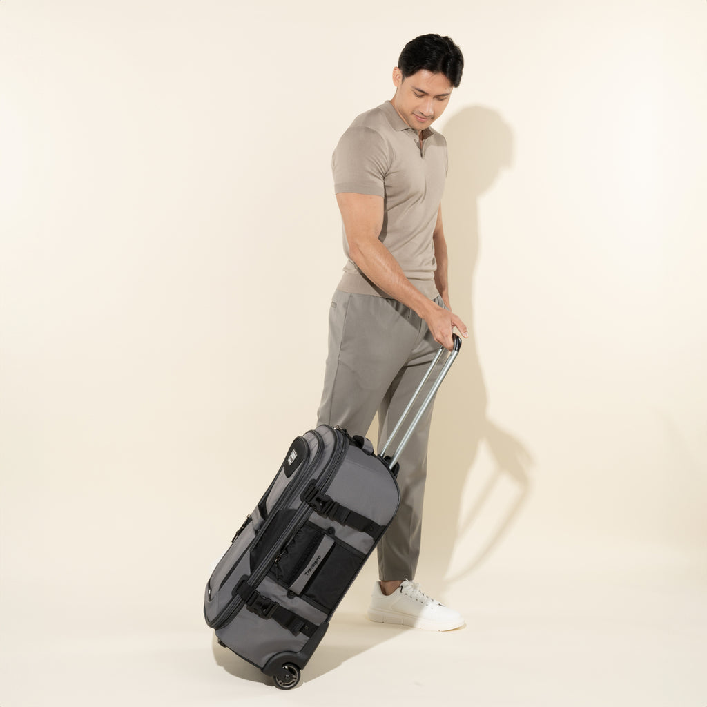 lifestyle view of a male model in a taupe polo shirt and grey pants tilting back and walking with a gray black bold by travelpro medium check-in expandable rollaboard suitcase, showing the bag in motion on its two wheels with the telescoping handle extended.