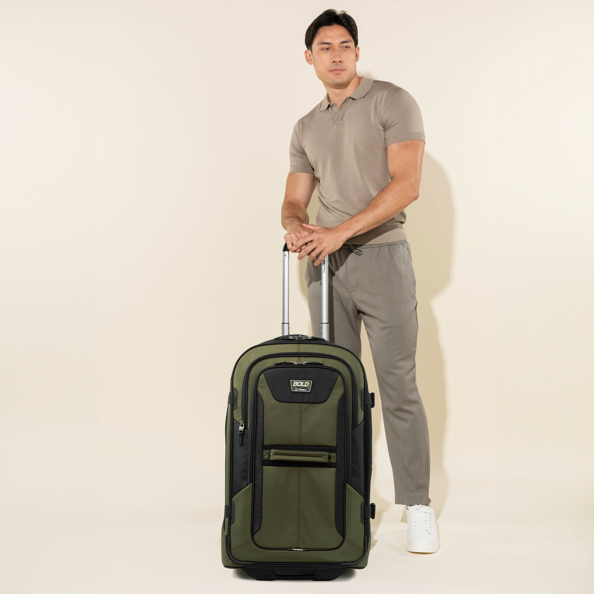 lifestyle view of a male model in a taupe polo shirt and matching trousers standing with his hands resting on the extended telescoping handle of an olive black bold by travelpro large check-in expandable rollaboard suitcase.