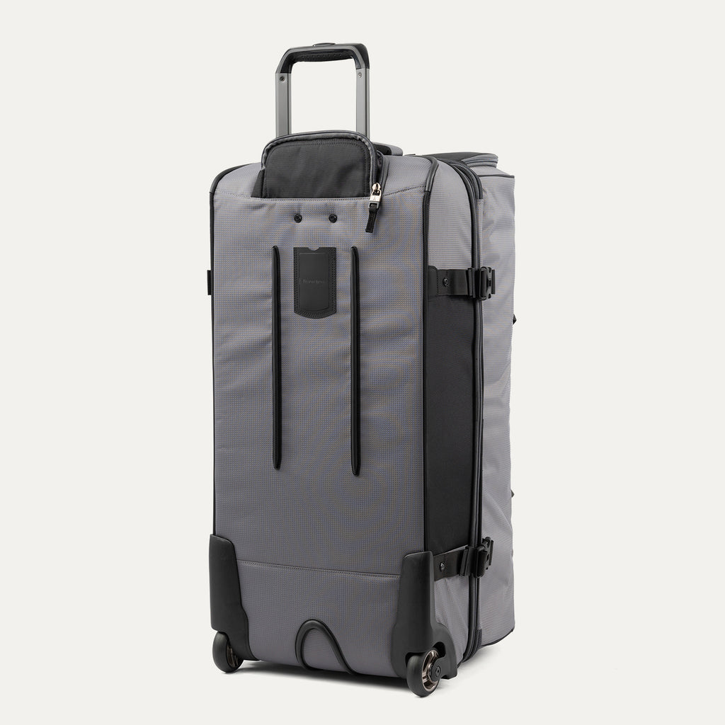 rear view of the gray black bold by travelpro large check-in drop-bottom rolling duffel bag standing vertically, showing its protective skid guards, reinforced wheel housings, two high-performance ball-bearing wheels, and extended telescoping handle.