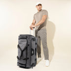 lifestyle view of a male model in a polo shirt and grey pants standing with the gray black bold by travelpro large check-in drop-bottom rolling duffel bag, showing its upright vertical scale and extended telescoping handle.