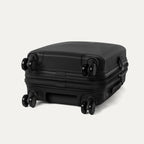 Side-profile view of the Optima Expandable Compact Carry-on Spinner in Black, highlighting the base construction and the self-aligning MagnaTrac® spinner wheels that ensure smooth, drift-free mobility.