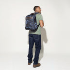 A person wearing a camouflage carry-on slim backpack with premium leather and polished chrome zippers, in a standing pose against a white background.