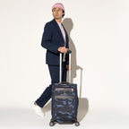 Person walking while rolling a Travelpro Platinum Elite Carry-On Spinner in camouflage using the extended PowerScope Extension Handle.
