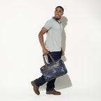 Person carrying the Travelpro Platinum Elite Underseat Tote in camouflage by the top handles, showcasing compact, on-the-go convenience.
