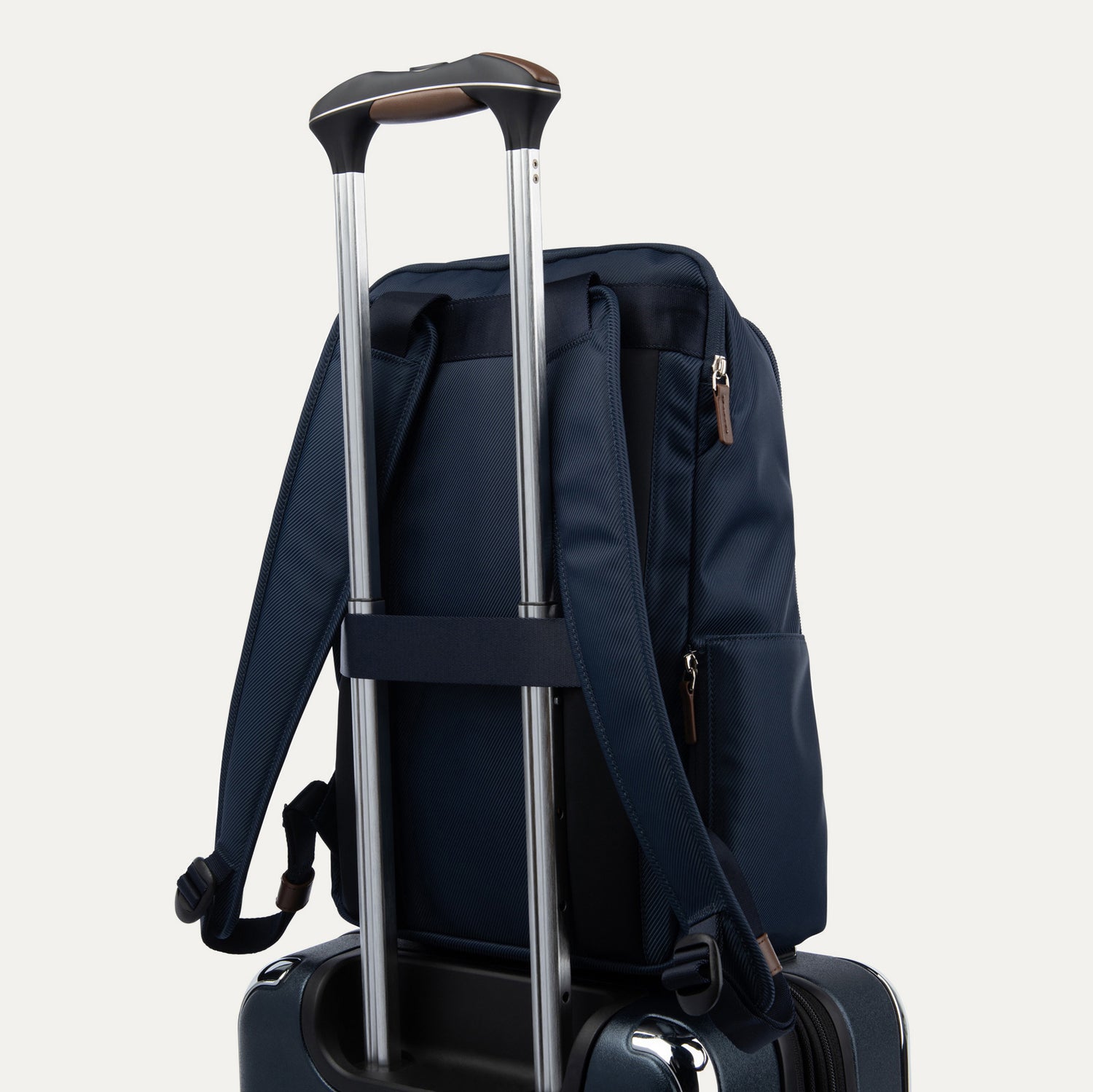 close-up rear view of the monaco blue travelpro x travel plus leisure slim backpack stacked securely on top of a suitcase using its built-in trolley sleeve, which slides over the extended aluminum telescoping handle featuring an elegant brown leather grip.