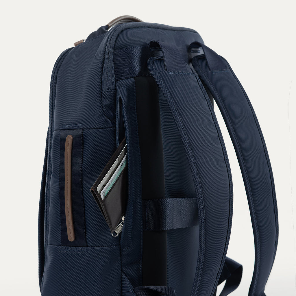 close-up side view of the monaco blue travelpro x travel plus leisure slim backpack, highlighting a zippered side storage pocket with an elegant brown leather trim accent, featuring a small personal item stored inside and durable navy blue fabric construction.