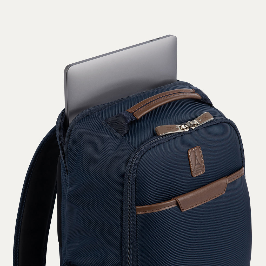 close-up side view of the monaco blue travelpro x travel plus leisure slim backpack, showcasing a silver laptop computer being slid into its dedicated rear zippered tech compartment, highlighting the rich blue fabric, padded shoulder strap, and elegant brown leather top carry handle.