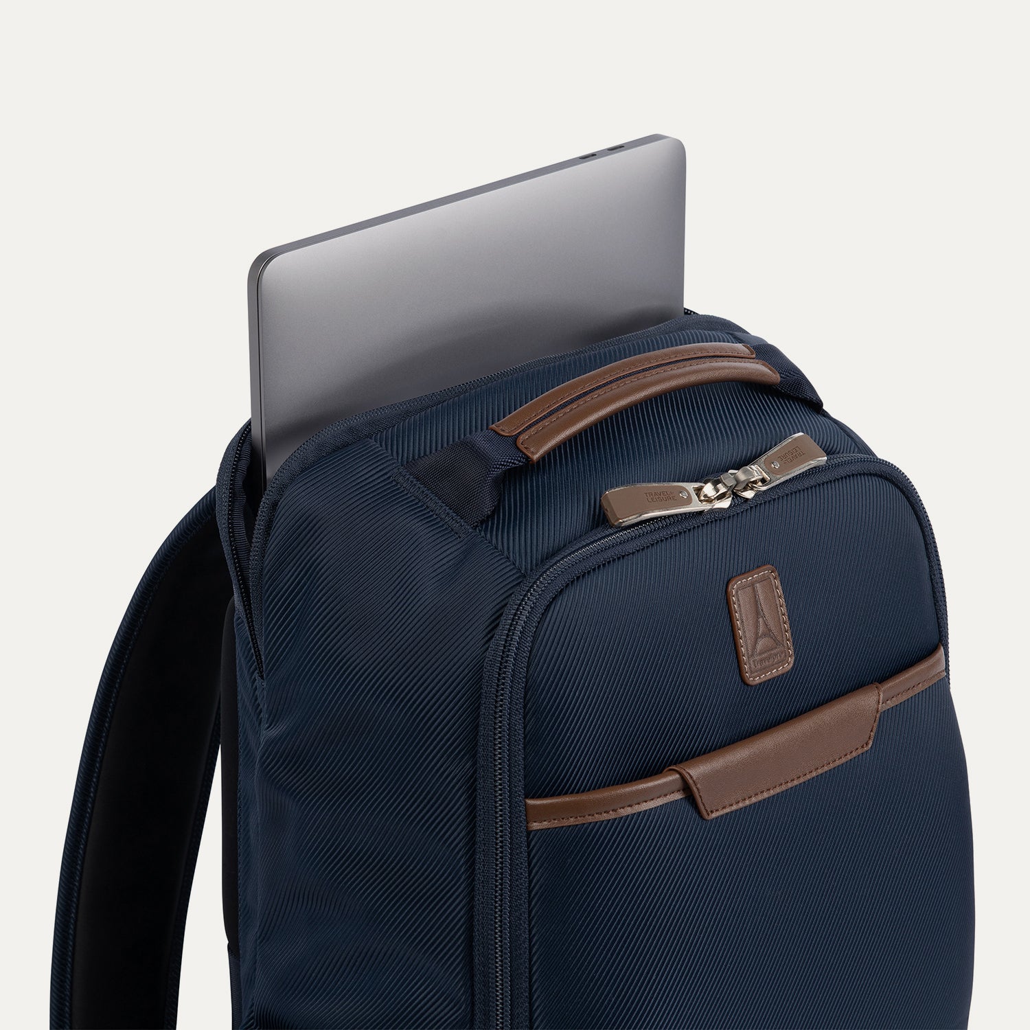 close-up side view of the monaco blue travelpro x travel plus leisure slim backpack, showcasing a silver laptop computer being slid into its dedicated rear zippered tech compartment, highlighting the rich blue fabric, padded shoulder strap, and elegant brown leather top carry handle.