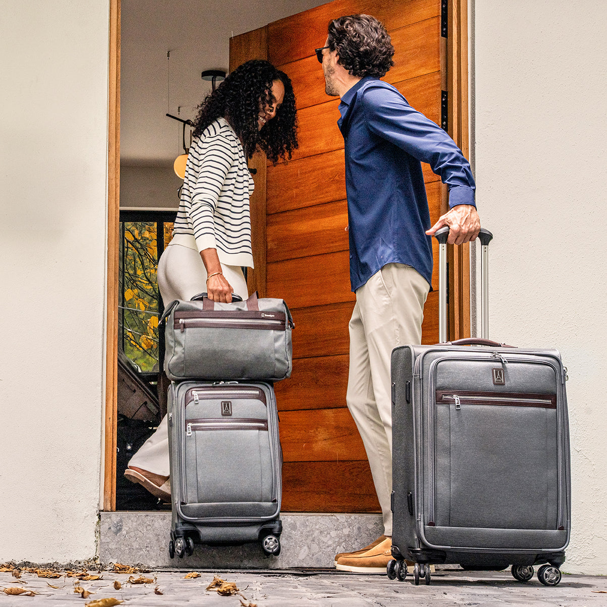 Two people with gray suitcases and a gray tote bag entering a house.