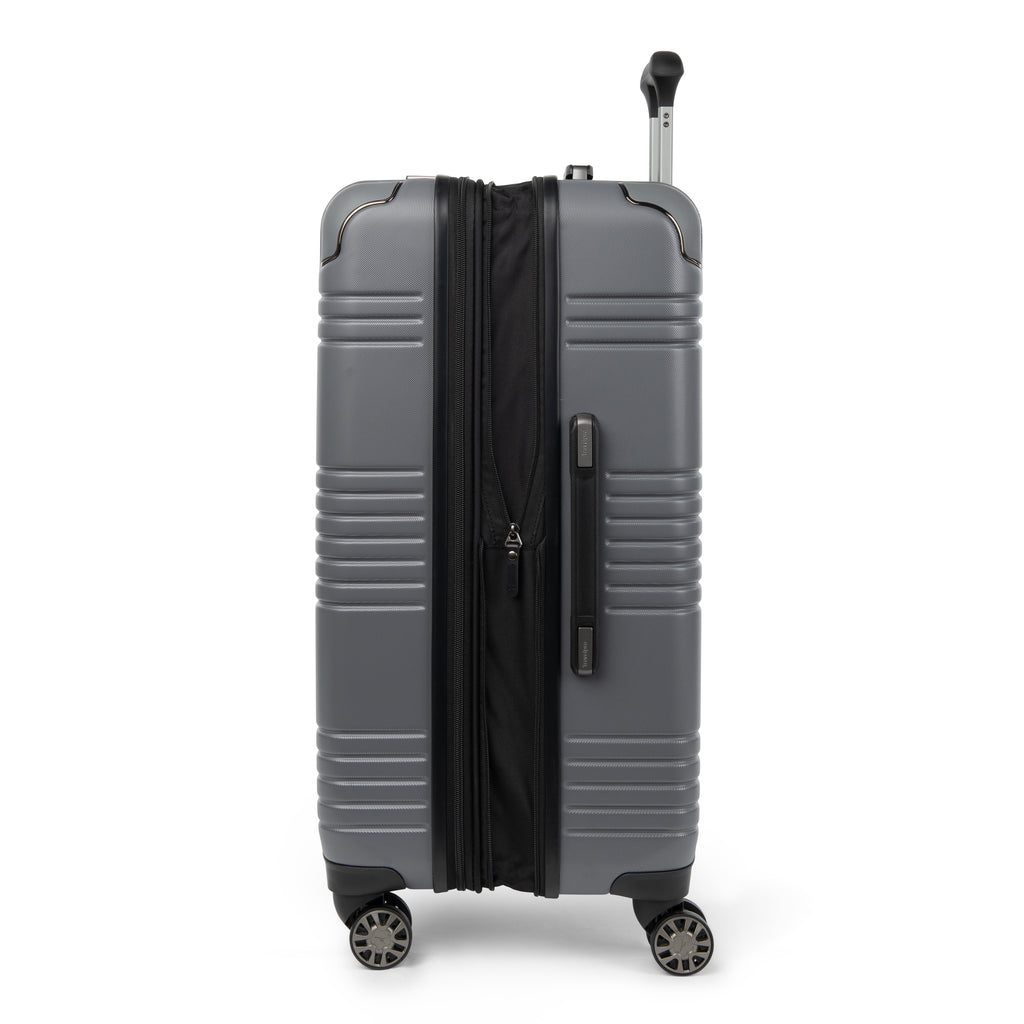 Side view of Travelpro Roundtrip Hardshell Medium Check‑In Suitcase in Gunmetal Gray, showing textured hardshell design, expansion zipper open, black telescoping handle, and four spinner wheels against white background.