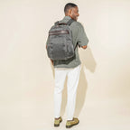 rear lifestyle view of a male model wearing the vintage grey travelpro platinum elite business laptop backpack 27l, showing the bag's fit against a casual olive green shirt and highlighting the dark brown leather trim and padded shoulder straps.