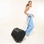 lifestyle view of a female model in blue trousers walking with the shadow black travelpro platinum® elite carry-on rolling garment bag, demonstrating its compact size and the height of the extended powerscope handle.