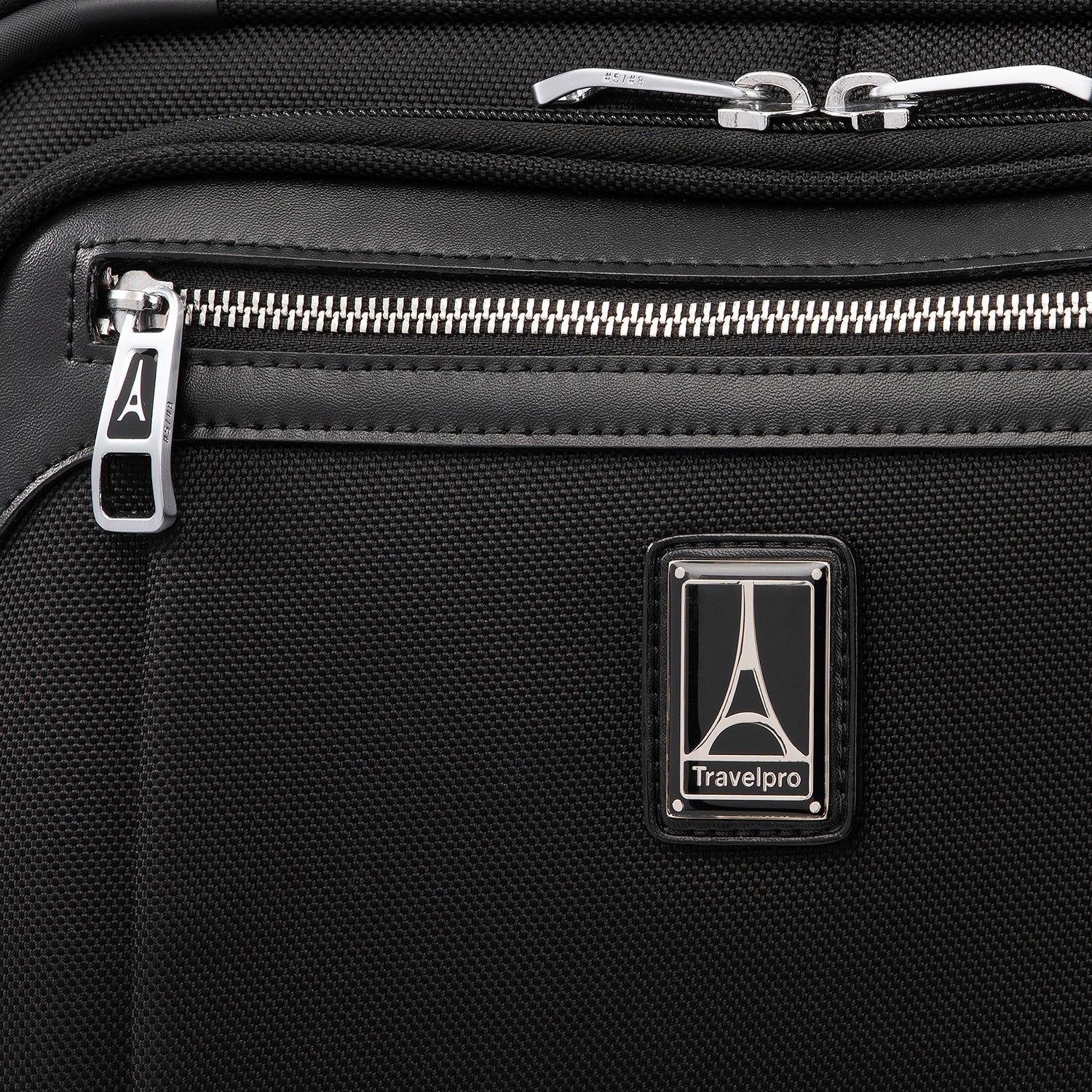 close-up view of the shadow black travelpro platinum® elite carry-on spinner tote, showing the chrome zipper pulls with the travelpro logo, a leather-trimmed pocket edge, and the metal travelpro logo badge on the front panel.