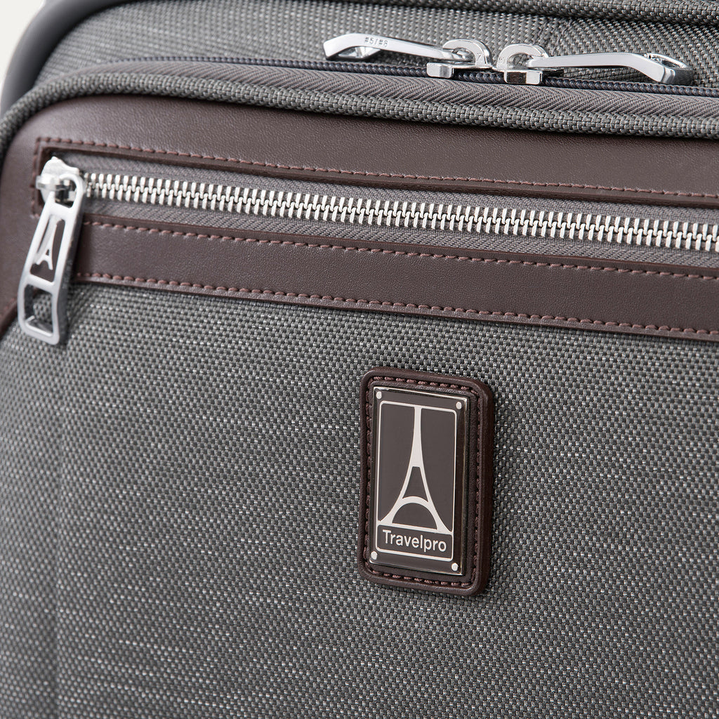 close-up view of the vintage grey travelpro platinum® elite carry-on spinner tote, showing the chrome zipper pulls with the travelpro logo, the brown leather-trimmed pocket edge, and the metal travelpro logo badge on the front panel.