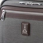 close-up view of the vintage grey travelpro platinum® elite carry-on spinner tote, showing the chrome zipper pulls with the travelpro logo, the brown leather-trimmed pocket edge, and the metal travelpro logo badge on the front panel.