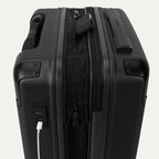 Close-up of the external charging ports on the Travelpro Platinum Elite Compact Carry-On Hardside Spinner in Shadow Black, showing a white cable plugged into the USB port and a power bank tucked into the dedicated battery sleeve within the expansion gusset.