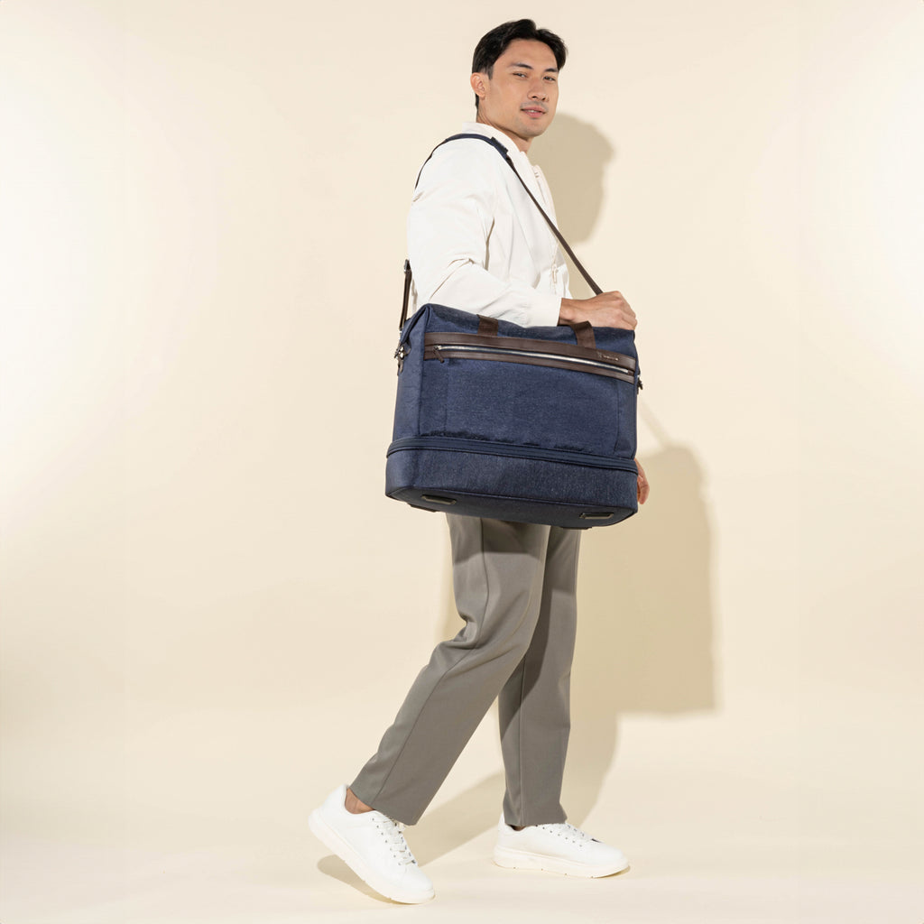 lifestyle shot of a male model walking while carrying the true navy travelpro platinum elite drop-bottom weekender bag using the adjustable shoulder strap, paired with casual white sneakers and tan trousers.