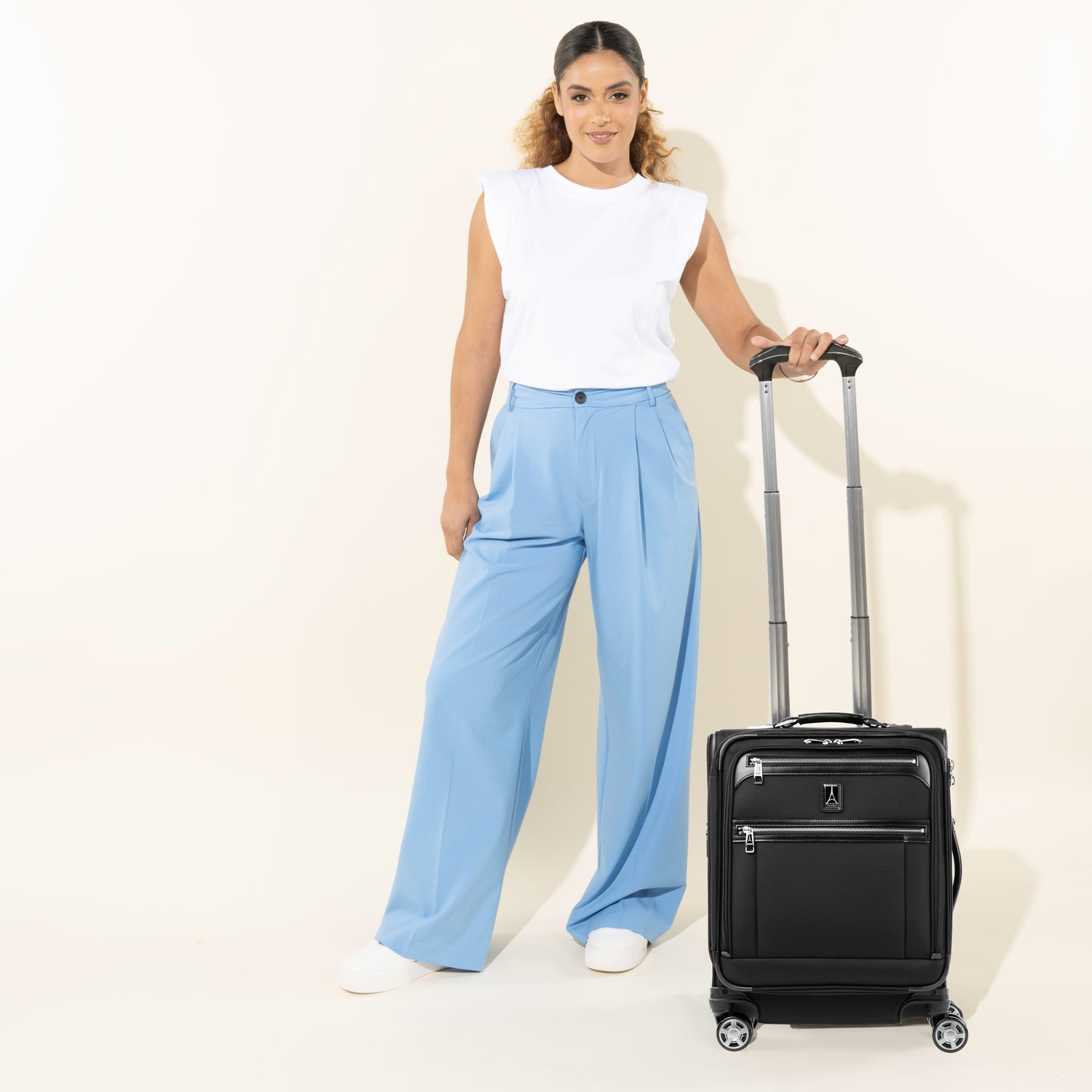 lifestyle view of a female model in light blue trousers and a white sleeveless top standing with the shadow black travelpro platinum® elite international carry-on spinner, showing the bag's height with the powerscope handle extended.