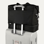 rear view of the shadow black travelpro platinum elite drop-bottom weekender bag, showing the integrated trolley sleeve securely stacked over the telescoping handle of a hardside suitcase for easy mobility.