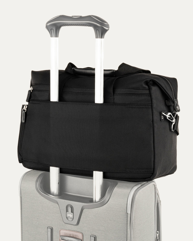 a black travelpro platinum elite underseat tote stacked on top of a silver rolling suitcase, with its integrated rear trolley strap securely slid over the luggage extension handle for easy transport.