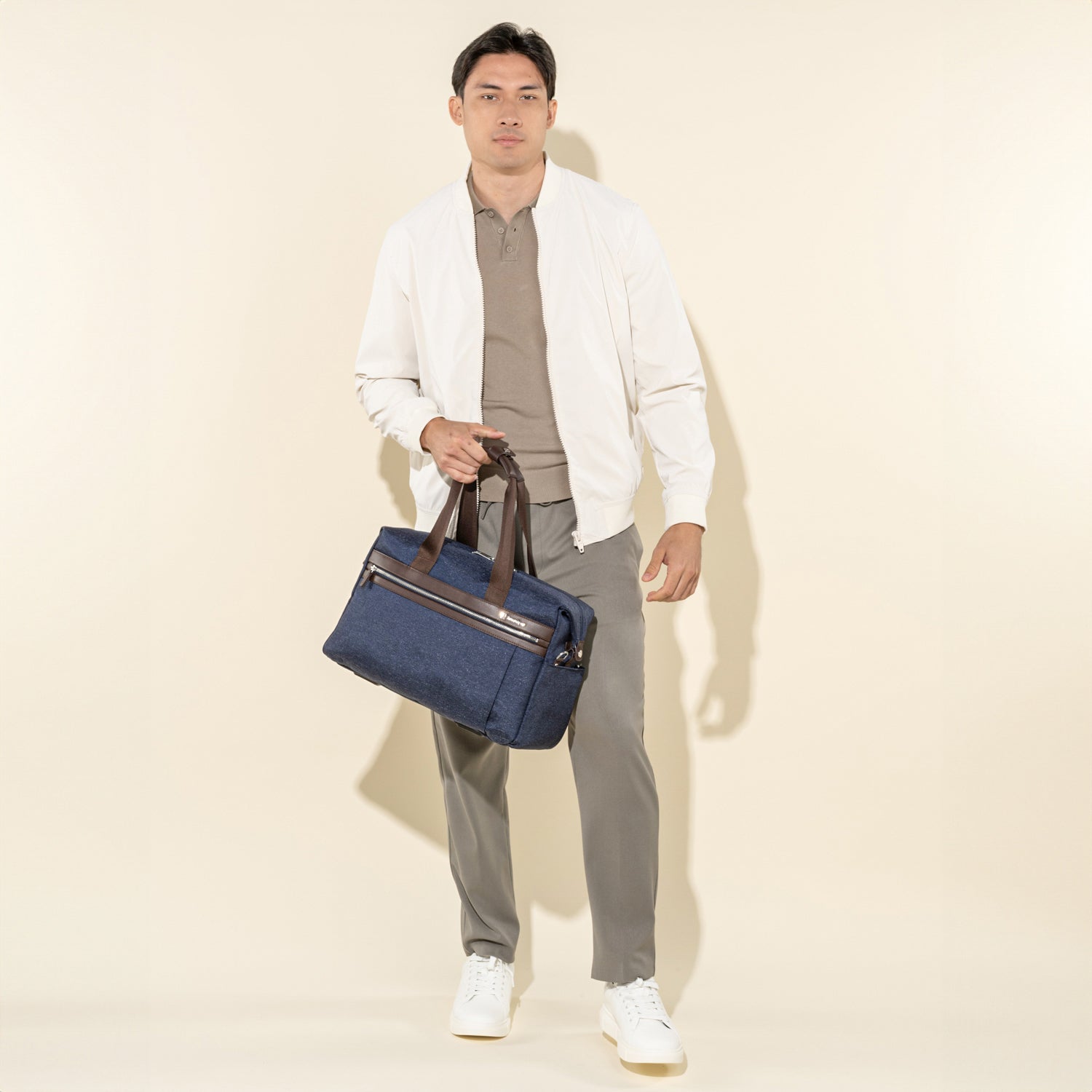 lifestyle shot of a male model in a white jacket and grey trousers, carrying a travelpro platinum elite underseat tote in true navy by its leather top handles.