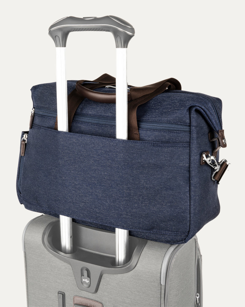 a true navy travelpro platinum elite underseat tote stacked on top of a silver rolling suitcase, with its integrated rear trolley strap securely slid over the luggage extension handle for easy transport.