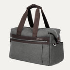 a three-quarter perspective view of the vintage grey travelpro platinum elite underseat tote, highlighting the textured grey fabric, brown leather trim, front zippered pocket, and side accessory pocket.